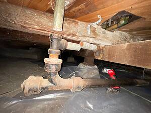 Rusty pipes and wooden joists in a dark crawlspace, with plastic vapor barrier on the ground and some metal brackets nearby.
