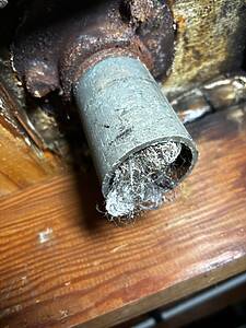 Close-up of a rusted pipe joint with brown corroded metal and frayed fiber insulation protruding from a worn sleeve.