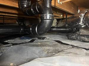 PVC plumbing pipes and joints running under a wooden floor joist, with foil-backed ducting nearby in a crawl space