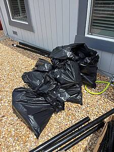Black trash bags piled on a gravel surface beside a blue-gray house wall with windows and scattered pipes nearby.