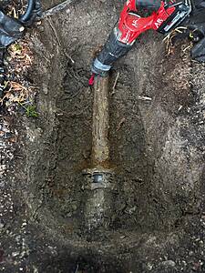 Rusty metal pipe buried in a muddy trench, with a red cordless drill resting near the surface.