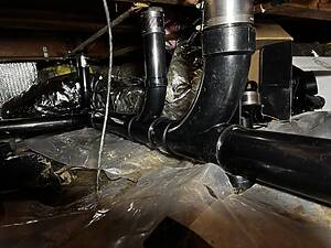 Basement crawl space with exposed HVAC ducts and black plastic pipes among foil insulation and scattered cardboard boxes.