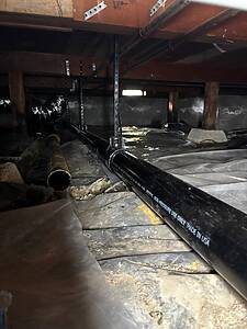 Crawlspace with exposed black PVC pipe labeled 'Non-Pressure Use Only' running along the floor, wooden joists above, and plastic sheeting on the ground.