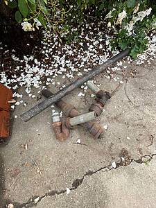 Rusty metal pipes lying on a concrete walkway beside a hedge with white flower petals scattered around.