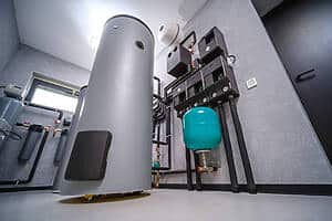 HVAC boiler room with a tall gray cylindrical tank, numerous pipes, and a teal expansion tank mounted on the wall.